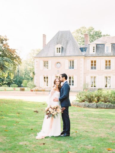 Bride and Groom smiling at each other at a Chateau de Bouthonvilliers Wedding. Bride is in a Alon Livne White gown and holding the bouquet down at her side. Groom is in a navy Chris Von Marital Suit.