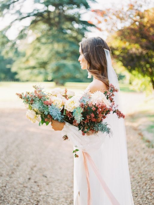 Bride looking off in the distance while holding her large floral bouquet with a soft pink silk ribbon flowing in the wind at Chateau de Bouthonvilliers Wedding