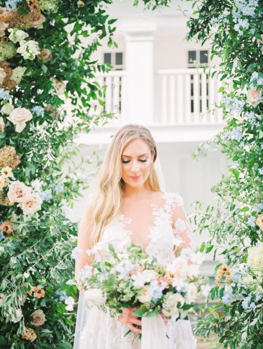 Bride with soft glam makeup and loose curls looking down at her large bouquet under a floral arch during a summer wedding at Zingerman's Cornman Farms Wedding Venue
