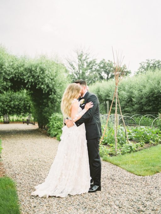 Bride in a Lace, Long Sleeve Galia Lahav Gown embracing with the Groom in a Black Tux at the gardens of Zingerman Cornman Farms Wedding